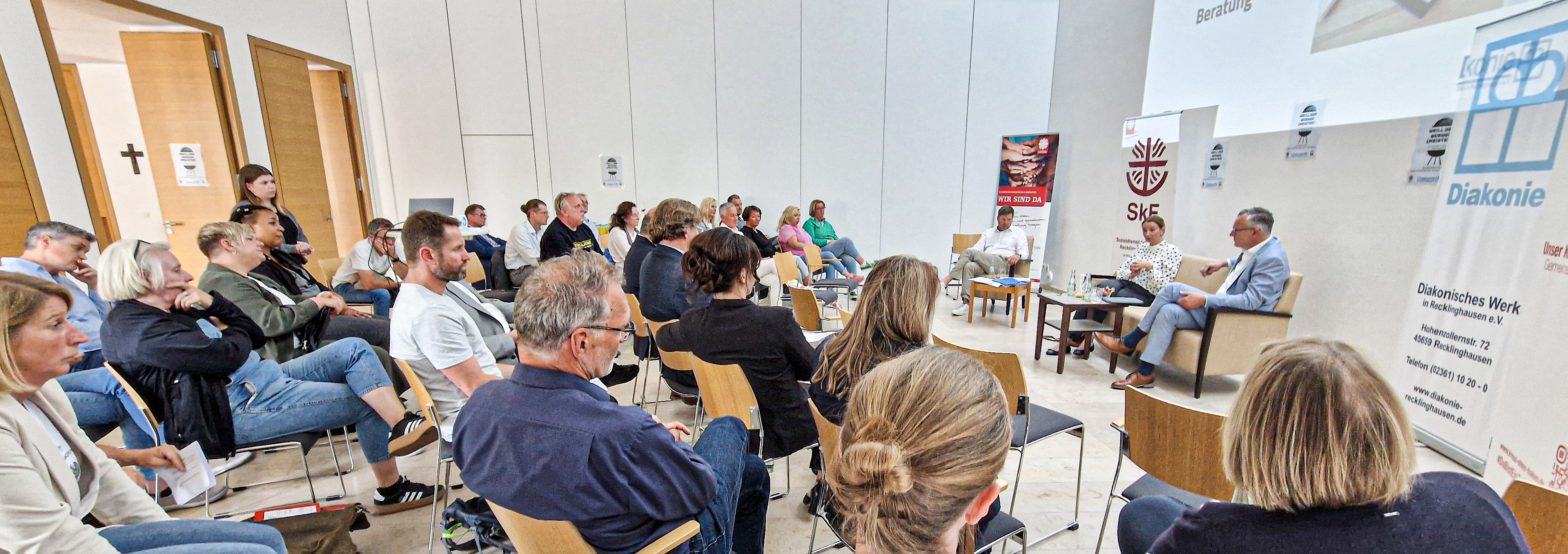 Mitarbeitende von SKF, Caritas und Diakone sitzen im Halbrund um das Podium mit Anna-Christian Rex, Axel Tschersich und Moderator Holger Boehnert.